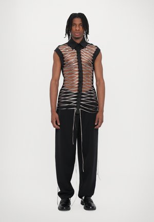 Schwarzes ärmelloses Shirt mit einem transparenten, überkreuzten Muster; Druckknopfverschluss vorne. Kombiniert mit schwarzen, locker sitzenden Hosen und Schuhen.