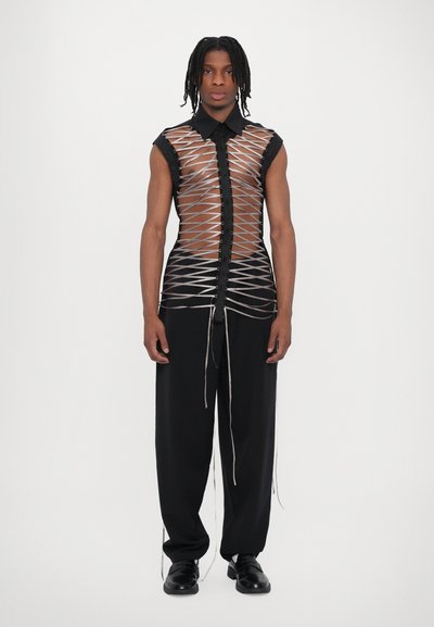 Schwarzes ärmelloses Shirt mit einem transparenten, überkreuzten Muster; Druckknopfverschluss vorne. Kombiniert mit schwarzen, locker sitzenden Hosen und Schuhen.