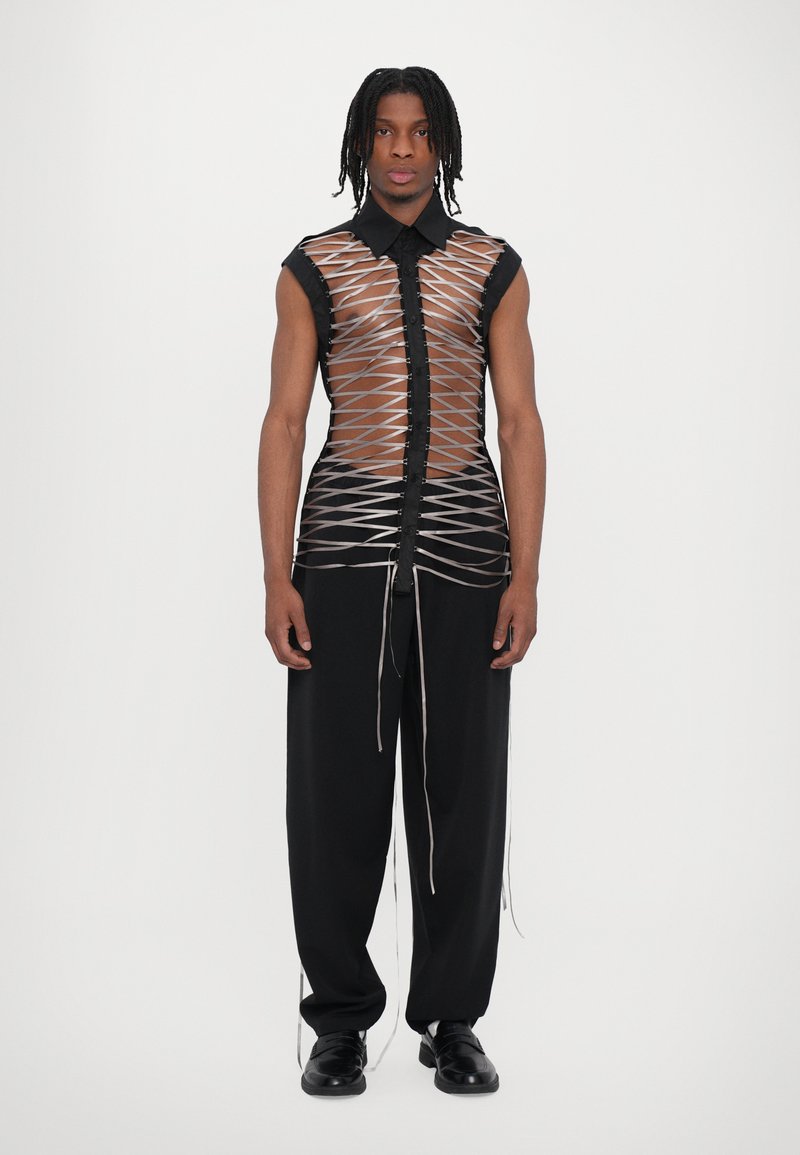 Chemise sans manches noire avec un motif transparent en croix ; fermeture à boutons à l'avant. Associée à un pantalon noir ample et des chaussures.