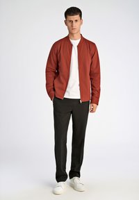 Röd bomberjacka över en vit t-shirt, svarta byxor och vita sneakers. Jackan har dragkedja och ribbade muddar, med en slät yta.