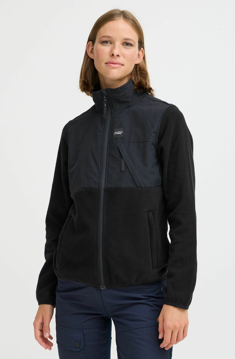 Juoda zip-up striukė su flisinėmis apatinių dalių, lygiu viršutiniu audiniu, šoninėmis kišenėmis ir mažu logotipu ant krūtinės.