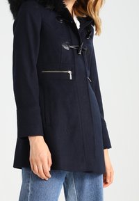 Navy blue coat with a faux fur hood, toggle and zipper closures, side pockets, and a fitted waist, made from a wool blend fabric.