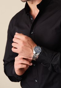 Silverfärgad metallklocka med en mörkblå cirkelformad urtavla, tre kronografunderklockor och ett armband av rostfritt stål, bärs på ett manligt handled.