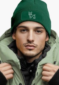 Green ribbed knit beanie with a rectangular patch featuring a white outline of a dog. Worn with a layered puffer jacket.