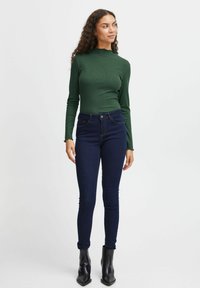 Grön ribbade långärmad tröja med vågiga kanter, ihop med mörkblå tight jeans och svarta ankelkängor, stående mot en neutral bakgrund.