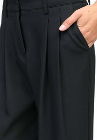 Main dans la poche droite d'un pantalon noir sur mesure avec des plis à l'avant et des passants de ceinture.