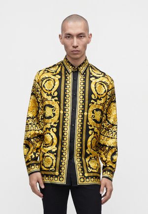 Uomo con testa rasata che indossa una camicia nera e dorata con motivi ornamentali a maniche lunghe, in piedi davanti a uno sfondo bianco uniforme.