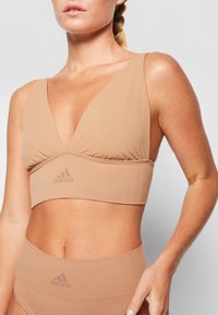 Bežinis sportinis liemenukas su giliu V formos iškirpimu, surinkta centrinė siūlė ir platus apatinis kraštas su subtiliu Adidas logotipu. Lygus, elastingas audinys.