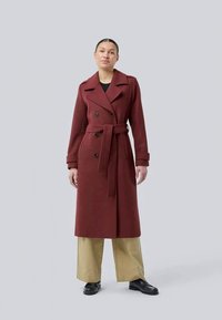 Trench coat bordeaux de dupla face com cinto. Apresenta grandes botões pretos, um colarinho largo e mangas longas. Usado sobre calças neutras.