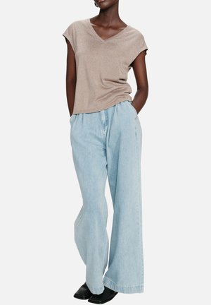 Femme portant un haut beige à manches courtes et col en V, un jean bleu clair à jambes larges avec des chaussures noires, mains dans les poches sur fond blanc.