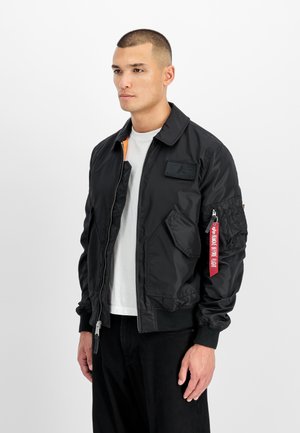 Schwarze Bomberjacke mit Reißverschluss vorne, gerippten Bündchen und einem Aufnäher auf der Brust. Verfügt über eine Reißverschlusstasche am Ärmel mit rotem Etikett-Detail.