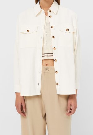 Kvinde iført en kremfarvet oversized skjorte med knapper over en cremefarvet strikket cropped top og højtaljede beige bukser med en stor knap.