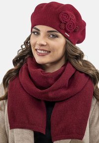 Berretto lavorato a maglia color borgogna con tre rose di tessuto e sciarpa dello stesso colore, caratterizzati da una morbida texture e uno stile drappeggiato.