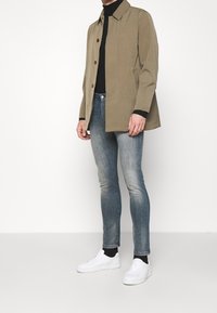 Muž oblečený v béžovém sakě na knoflíky přes černý rolák, vybledlých skinny džínech, černých ponožkách a bílých teniskách, stojící na bílém pozadí.