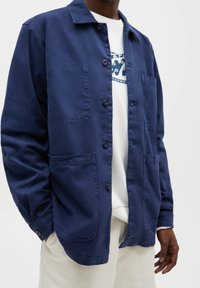 Mörkblå bomullsjacka med knappstängning fram, två bröstfickor och avslappnad passform. Den har en klassisk krage och kontrasterande knappar.