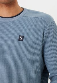 Lichtblauwe gebreide trui met een textuurpatroon, met een ronde halslijn en een klein zwart logo-embleem op de borst.