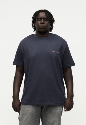 Sort mand med korte dreadlocks iført en mørkeblå T-shirt med lille "originals" tekst på brystet og grå jeans, stående mod en simpel baggrund.