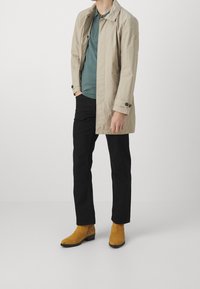 Beige keve trench coat yllä teal väristä polo-paitaa, yhdessä mustien housujen ja keltaisten mokka nilkkasaappaiden kanssa. Siisti, minimalistinen muotoilu.