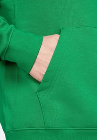 Sweat-shirt vert avec une poche avant, des poignets côtelés et une texture douce, mis en valeur par un gros plan de la main reposant dans la poche.