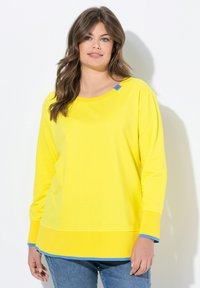 Chemise jaune à manches longues avec un col rond, des poignets et un ourlet côtelés, présentant des accents bleus au col et aux poignets. Texture douce et lisse.
