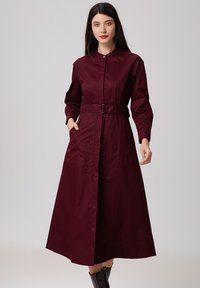 Robe à manches longues bordeaux avec des boutons, dotée d'une taille ceinturée, de poches latérales et d'une silhouette évasée. Matériau en coton doux.