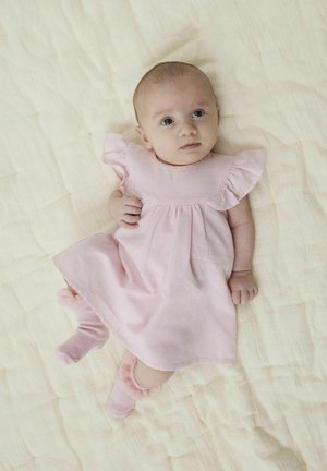 Vestido de algodón rosa claro con mangas acampanadas, combinado con calcetines rosa a juego con volantes, sobre un suave fondo beige texturizado.