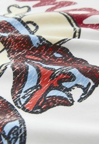 Közelkép egy szövetről, melynek színes, absztrakt mintája élénk piros, kék és fekete formákat ábrázol fehér háttéren.