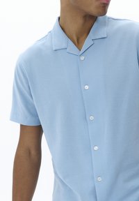 Camicia a maniche corte con colletto in tessuto azzurro chiaro; presenta una chiusura frontale con cinque bottoni bianchi e una superficie liscia.