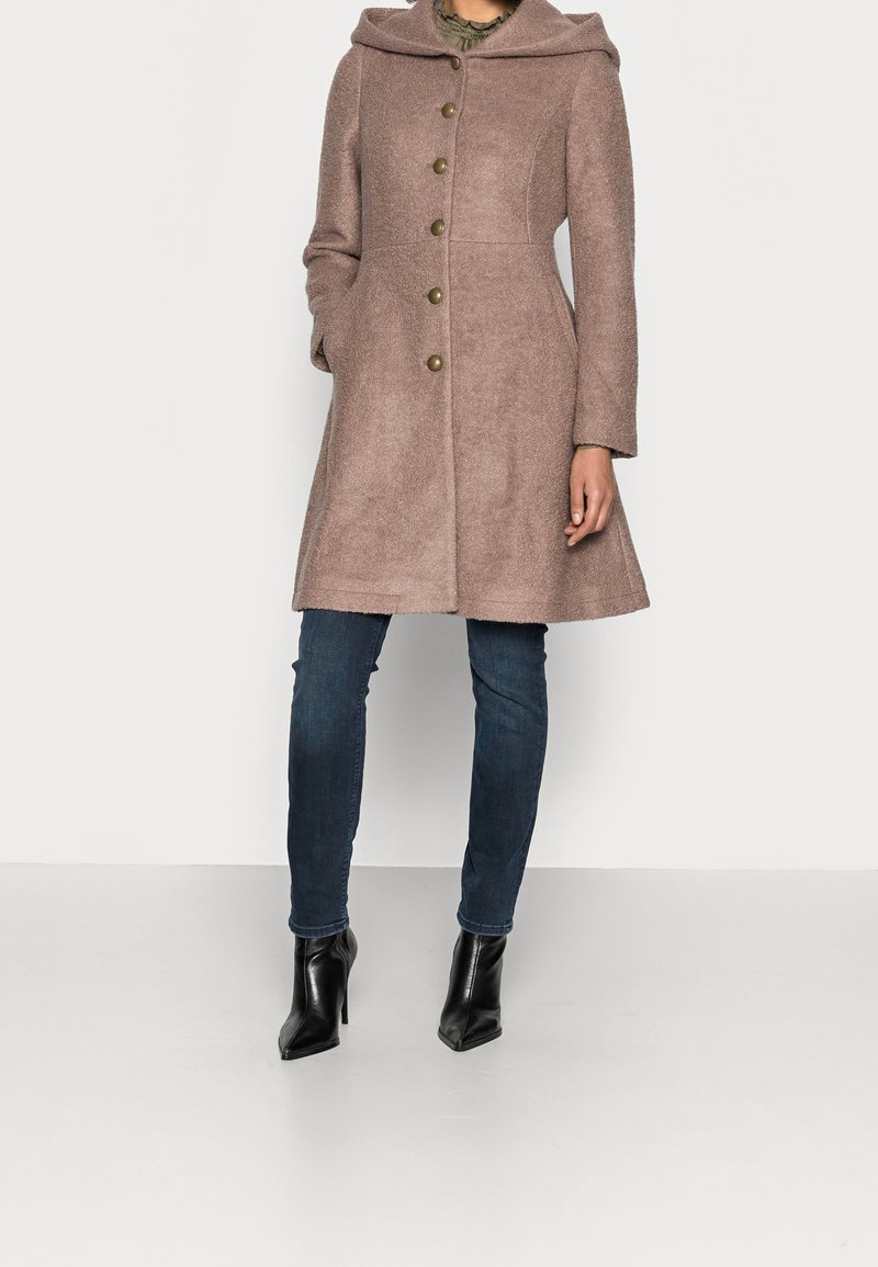 Femme portant un manteau en laine marron à capuche longueur genou avec des boutons, un jean bleu foncé et des bottines noires à talons, debout devant un fond clair.