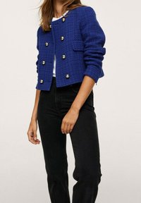 Veste bleue texturée avec boutons dorés, coupe courte, encolure ronde et manches longues. Associée à un jean noir.