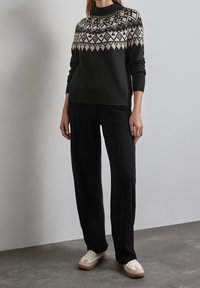 Svart stickad tröja med geometriskt mönster i krämfärg, rund halsringning och randiga muddar, tillsammans med svarta byxor och beige sneakers.