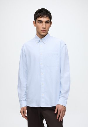 Jeune homme portant une chemise bleu clair boutonnée avec une poche poitrine et un pantalon sombre, debout devant un fond blanc uni.