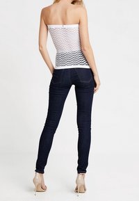 Valkoinen olkaimeton toppi, jossa on timanttimainen verkkomuotoilu, yhdistettynä tummansinisiin tiukkoihin farkkuihin ja nude-väriin korkokenkiin.