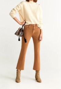 Pull en maille crème avec des boutons aux épaules, associé à un jean évasé marron. Les accessoires incluent un sac à motifs et des bottes cheville beiges.