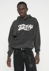 Hall hõlmikuga hall dressipluus, mille ees on kulunud valge graafiline logo, kombineeritud mustade teksadega, millel on naelutatud detailid ja narmendavad servad.