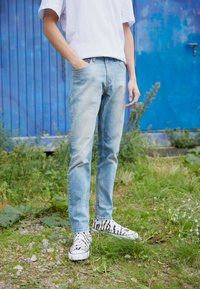 Light blue denim jeans with a slim fit, paired with white sneakers featuring a black zebra stripe pattern. Grass and foliage in background.