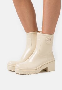 Beige rubberen enkellaarzen met een glanzende afwerking, een chunky hak en reliëf logo-detail. Kenmerken zijn een ronde neus en een gestructureerde zool.
