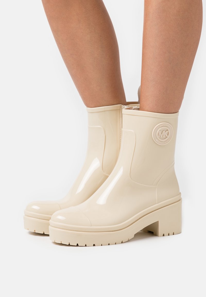 Beige rubberen enkellaarzen met een glanzende afwerking, een chunky hak en reliëf logo-detail. Kenmerken zijn een ronde neus en een gestructureerde zool.