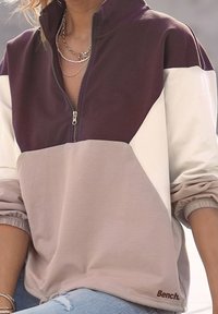 Sweter w kolorze burgunda, kremowym i jasnym beżu. Posiada półzapinaną dekolt, elastyczne mankiety oraz logo "Bench." na końcu. Gładka faktura materiału.