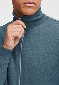 Sweat-shirt à col haut zippé en tissu bleu canard foncé, avec un col côtelé et une fermeture éclair argentée visible. Texture douce avec une silhouette ajustée.