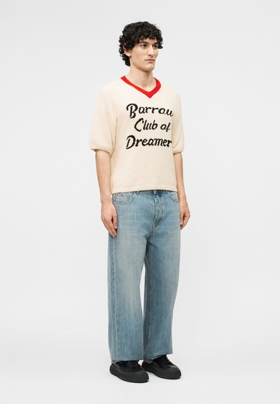 Jeune homme debout portant un pull à manches courtes crème avec un col rouge et le texte « Barrow Club of Dreamers », un jean bleu clair et des baskets noires.