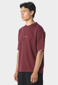 Burgundské tričko s krátkým rukávem vyrobené z jemného bavlněného materiálu, s kulatým výstřihem a vyšívaným textem na hrudi, s malým logem na rukávu.