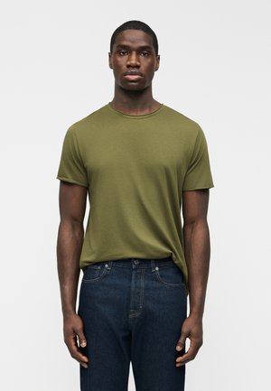 Muž v olivovo zelenej tričku s krátkym rukávom a okrúhlym výstrihom, mierne zastrčenom do tmavomodrých džínov, stojaci pred jednoduchým svetlým pozadím.