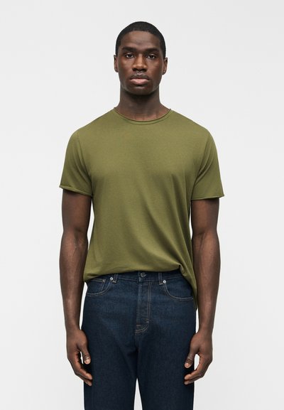 Man wearing olive green short-sleeve crew neck t-shirt tucked slightly into dark blue jeans, standing against a plain light background.