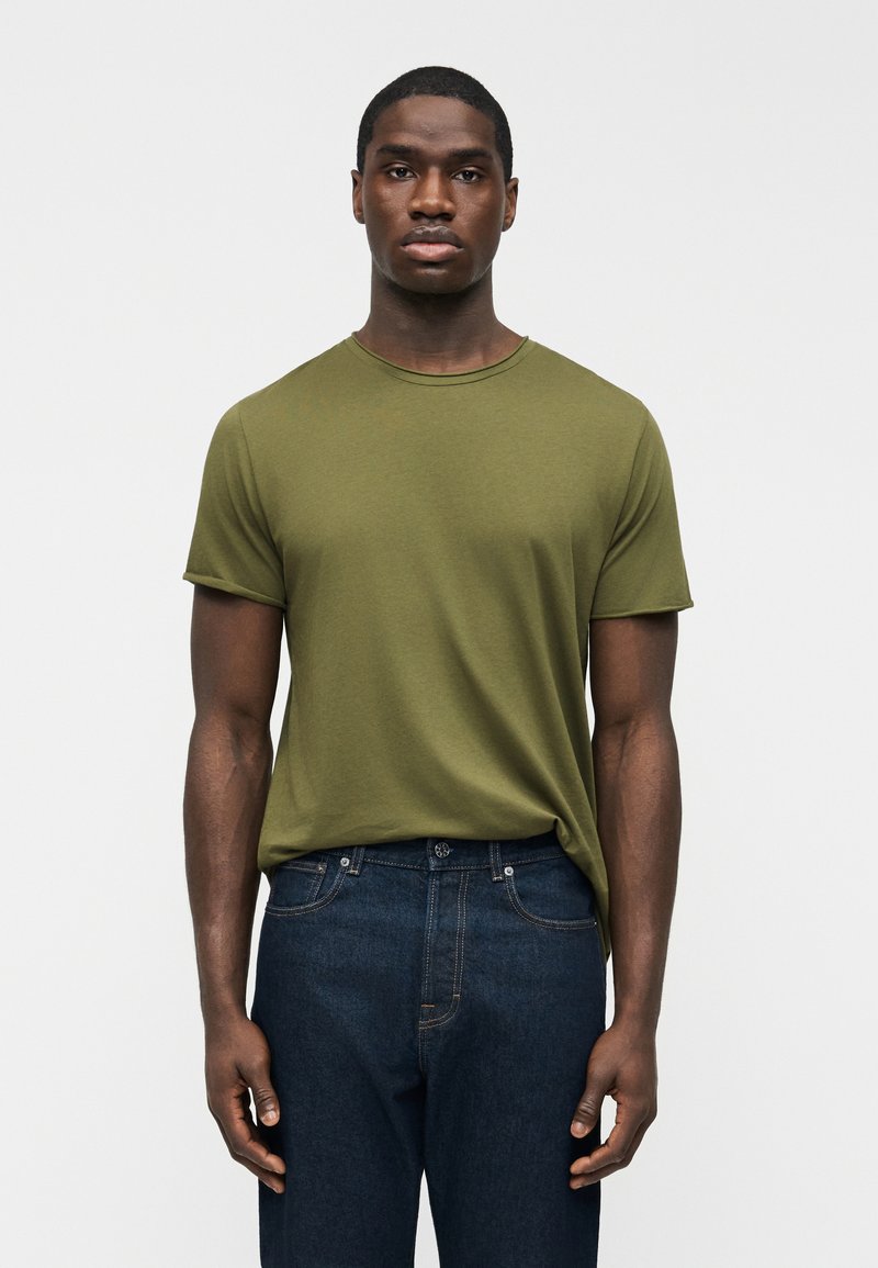 Άνδρας που φοράει ελαιί πράσινο κοντομάνικο μπλουζάκι με στρογγυλή λαιμόκοψη, ελαφρώς μισοβαμμένο μέσα σε σκούρα μπλε τζιν, στέκεται μπροστά σε ένα απλό φωτεινό φόντο.