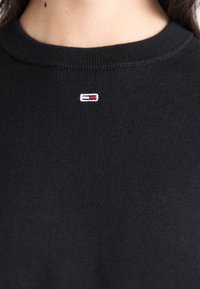 Pull en maille noir avec un col rond et un petit logo brodé en rouge, blanc et bleu sur la poitrine.