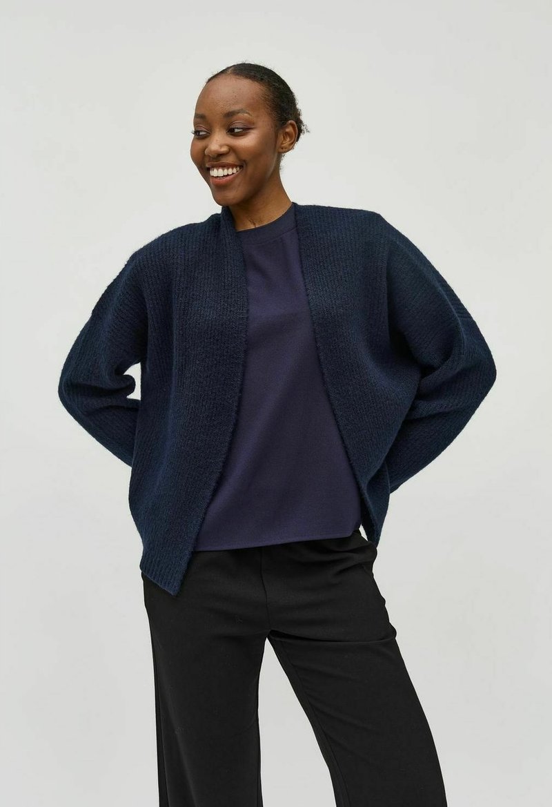 Cárdigan de punto azul marino con abertura frontal, corte relajado y mangas anchas; combinado con una blusa a juego y pantalones negros.