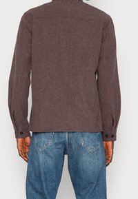 Hnědá košile s dlouhým rukávem z texturované látky, rovného střihu, se zapínáním na knoflíky na manžetách a klasickým límečkem, ve spojení s modrými džínami.