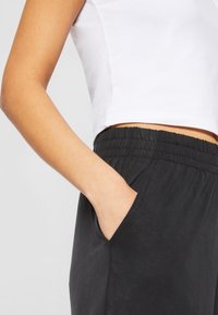 Gros plan sur le torse d'une personne portant un top court blanc et un pantalon noir taille élastique, avec une main posée négligemment dans la poche latérale.