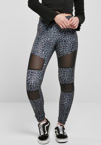 Leopardmønster leggings i grå og sort, med gjennomsiktige mesh-utskjæringer ved knærne, laget av elastisk stoff for komfort. Svart sneakers.